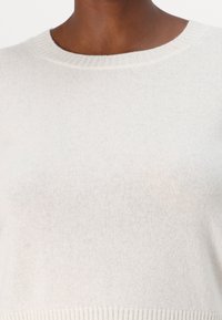 Gros plan d'une personne portant un pull ras du cou tricoté beige clair avec col et ourlet côtelés.