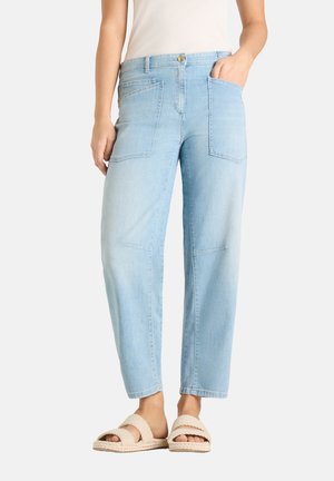 Light blue denim pants with large front pockets worn with beige braided sandals and a white top, standing with one hand in pocket.