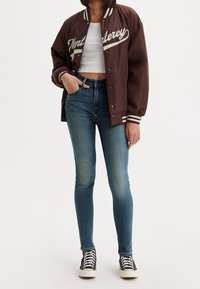 Chaqueta bomber marrón con letras crema, puños a rayas blancas, combinada con jeans de mezclilla azul ajustados y zapatillas negras de caña alta.