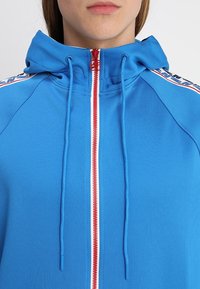 Sudadera con cremallera azul clara, con una cremallera roja, detalles blancos y cordones. Presenta una capucha y rayas laterales con detalle en texto.