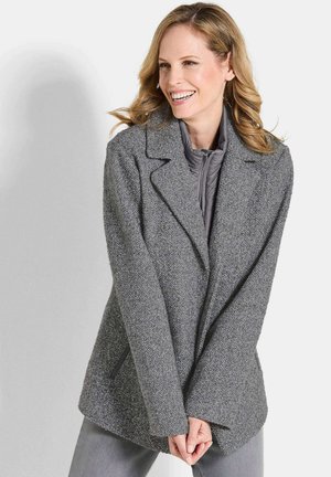 Femme souriante, portant un blazer gris texturé sur une veste zippée en quilt gris, se tenant les mains jointes devant elle, sur un fond clair.