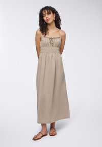 Abito maxi senza maniche in tessuto beige chiaro, con vita arricciata, scollatura arricciata e dettagli con bottoni sul davanti. Abbinato a sandali marroni.