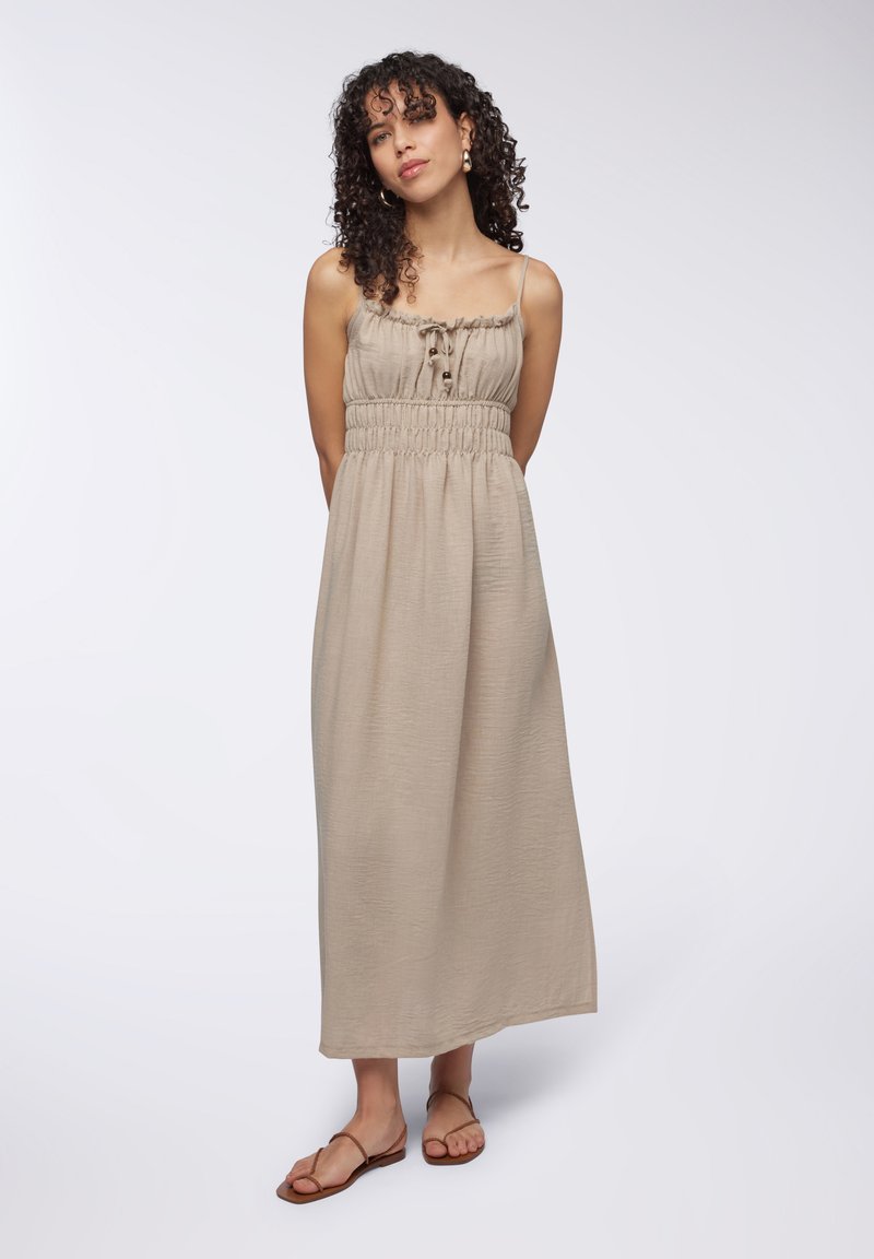 Abito maxi senza maniche in tessuto beige chiaro, con vita arricciata, scollatura arricciata e dettagli con bottoni sul davanti. Abbinato a sandali marroni.