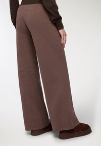 Kobieta w brązowych szerokich spodniaczh teksturowych, ciemno-brązowym swetrze i brązowych butach na platformie stoi na tle jasnym.