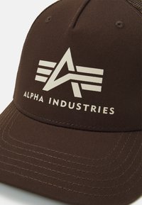 Casca de baseball maro cu logo-ul alb "Alpha Industries" care prezintă un "A" stilizat și dungi orizontale pe panoul din față.