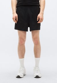 Μαύρο αθλητικό σορτς με ομαλή υφή, πλαϊνές τσέπες, ελαστική μέση και διακριτική λεπτομέρεια λογότυπου. Φοριέται με λευκά αθλητικά παπούτσια και κάλτσες.