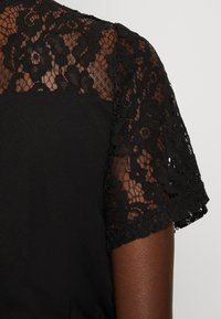 Robe noire avec des manches courtes en dentelle à motifs floraux, contrastant une texture transparente avec un corsage noir uni.