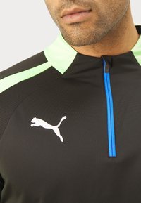 Svart träningsskjorta med gröna accenter, med en blå dragkedja och en vit logotyp. Tillverkad av texturerat tyg med en strukturerad design.