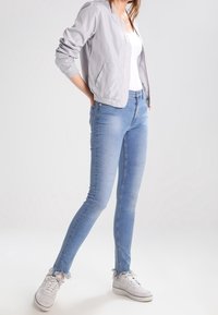 Veste bomber gris clair portée sur un débardeur blanc, associée à un jean skinny bleu clair avec des ourlets effilochés et des baskets blanches.