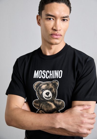 T-shirt en coton noir avec un grand graphique d'un ours et le texte "MOSCHINO" au-dessus du design. Le modèle croise les bras.