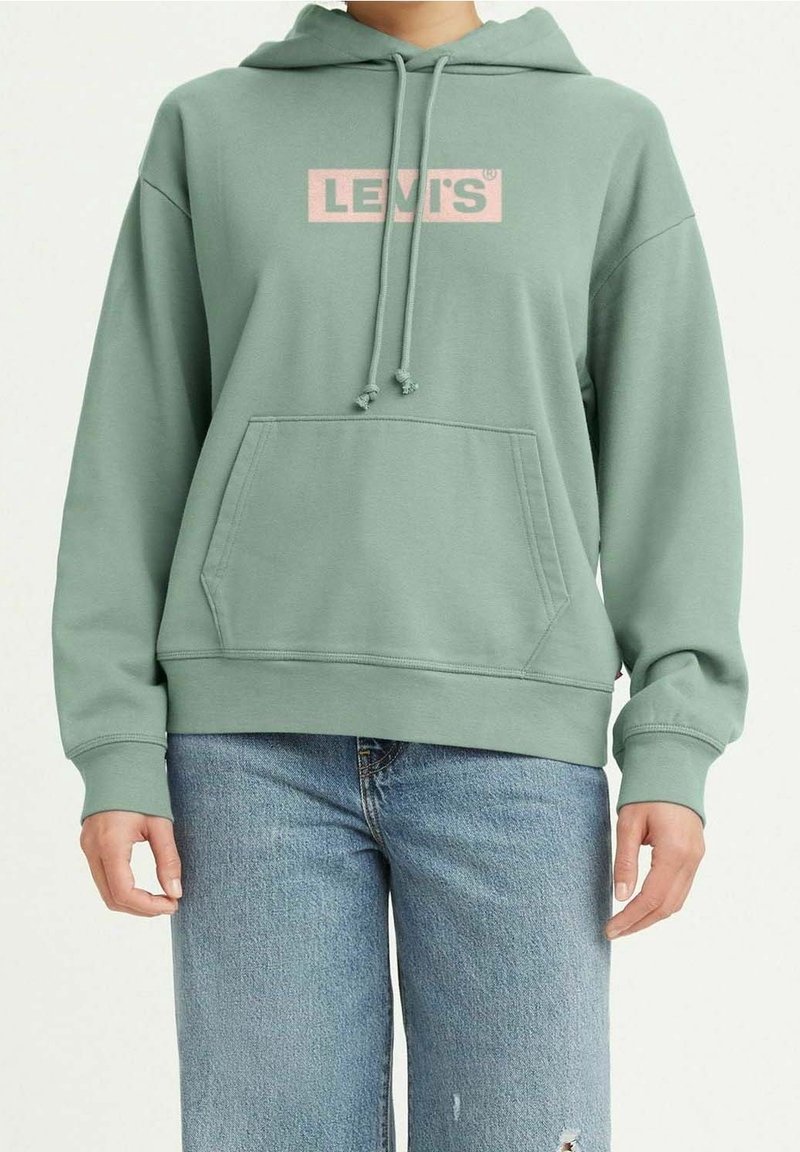Personne portant un sweat à capuche Levi's vert sauge avec poche avant et un jean bleu légèrement usé sur la cuisse.