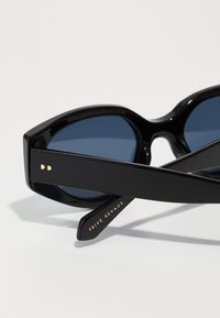 Óculos de sol pretos com um design geométrico ousado, lentes azuis e detalhes em dourado, apresentando o nome da marca "PRIVÉ REVAUX" na armação.