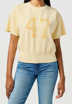 Persoon in een lichtgele kortekд mouwen sweatshirt met het nummer "47" in vervaagd geel en strakke blauwe spijkerbroek tegen een neutrale achtergrond.