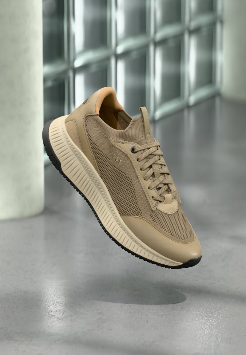Μπεζ ελαφρύ αθλητικό παπούτσι με λεπτομέρειες από δίχτυ και δέρμα, σχέδιο με κορδόνια, παχιά υφή στη σόλα, αιωρείται πάνω από γυαλισμένο δάπεδο από σκυρόδεμα σε εσωτερικό χώρο.