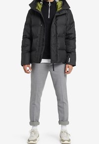 Musta toppatakki, jossa on vihreä vuori, korkea kaulus, vetoketjukiinnitys ja sivutaskut. Yhdistetty harmaisiin kapeneviin housuihin ja lenkkareihin.