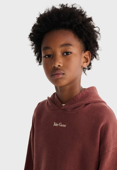 Junge Person mit lockigem Haar, die einen braunen Kapuzenpullover mit dem Text "Bobo Choses" trägt und direkt in die Kamera vor einem einfarbigen Hintergrund blickt.