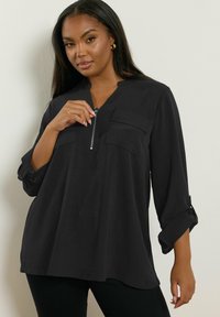 Maglietta nera a maniche lunghe con colletto zip, due tasche sul petto, vestibilità comoda e maniche arrotolate. Tessuto morbido e leggero.