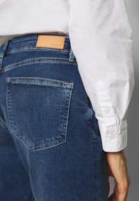 Tmavě modré džíny vyrobené z džínsoviny s hladkým povrchem, s kapsami na zadní straně a hnědou koženou nášivkou s názvem "S. OLIVER." Viditelný rukáv bílé košile.