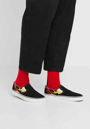 Voeten met zwarte slip-on schoenen met vlammenontwerp en felrode sokken, gecombineerd met zwarte cropped broeken op een eenvoudige vloer.