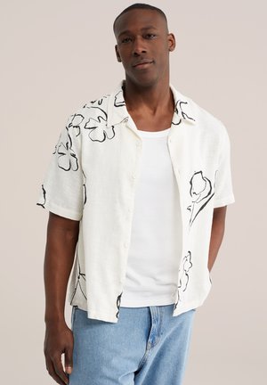 Mand iført en hvid kortærmet skjorte med knapper og sorte blomstermønstre over en hvid tanktop og lyseblå jeans.