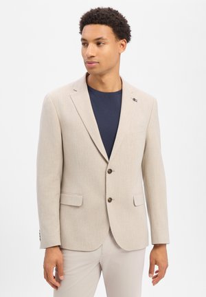 Man wearing beige blazer over navy shirt, beige pants, standing against plain white background, looking left.