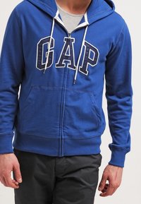 Uomo che indossa un hoodie blu con zip e logo "GAP" e lacci bianchi, abbinato a pantaloni neri e una maglietta grigia chiara sotto.