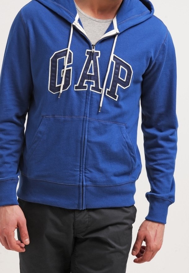 Uomo che indossa un hoodie blu con zip e logo "GAP" e lacci bianchi, abbinato a pantaloni neri e una maglietta grigia chiara sotto.