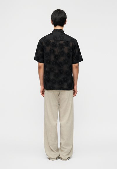 Άτομο με κοντά μαύρα μαλλιά που φορά διαφανές μαύρο κοντομάνικο πουκάμισο με λουλούδια και άνετο μπεζ παντελόνι, στέκεται γυρισμένο με την πλάτη σε απλό φόντο.