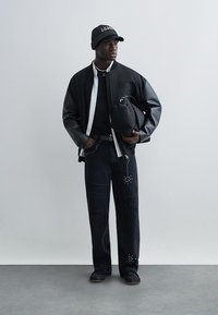 Homme portant une veste avec manches en cuir noir, un jean noir déchiré avec des clous, une casquette noire et tenant un sac matelassé noir.