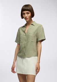 Camicia a maniche corte con colletto in morbido tessuto verde oliva testurizzato, dotata di due tasche sul petto. Abbinata a una gonna di jeans bianca.