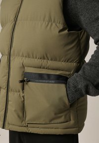 Olijfgroene gewatteerde vest met een ritszak en zwarte accenten. Glad, duurzaam materiaal met een gestikt textuur voor isolatie.