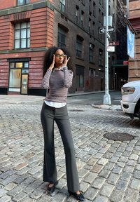 Črtasta majica z dolgimi rokavi in belim ovratnikom, črne hlače s flared krojem in črne čevlje z naborki, postavljeni pred ozadje opečne stavbe.