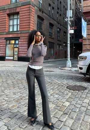 Haut à rayures à manches longues avec un col blanc, pantalon noir évasé, et chaussures noires avec volants, le tout sur fond d'un bâtiment en briques.