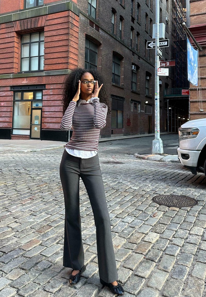 Črtasta majica z dolgimi rokavi in belim ovratnikom, črne hlače s flared krojem in črne čevlje z naborki, postavljeni pred ozadje opečne stavbe.