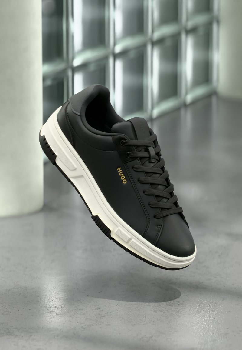 Baskets basses noires avec semelle blanche et logo "HUGO" doré sur le côté, flottant au-dessus d'un sol en béton poli.