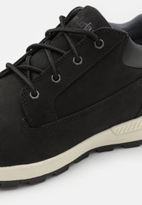 Svarta mockaskor med en strukturerad yta, utrustade med svarta snören, metallögon och en kontrasterande vit och svart gummisula.