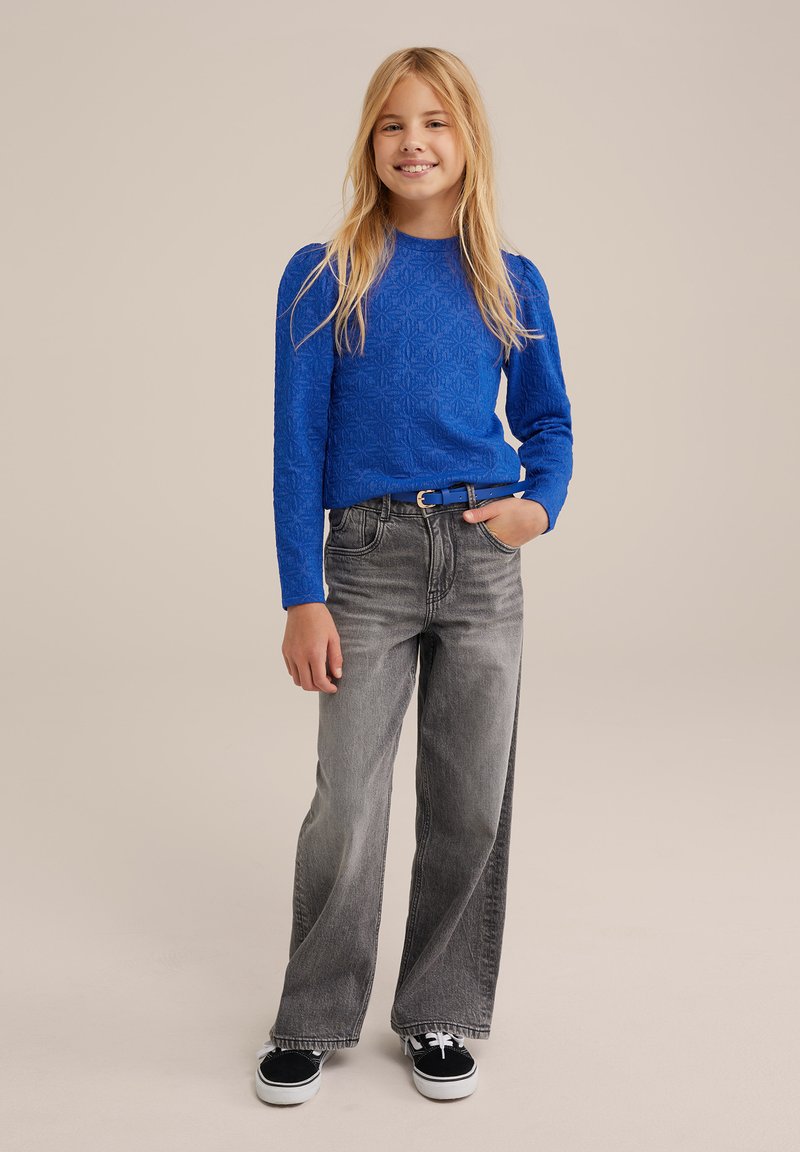 Blå texturerad långärmad topp, grå vida jeans och svarta sneakers. Modellen står med ena handen i fickan mot en neutral bakgrund.