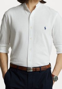 Camisa blanca de botonadura con cuello mao, con un pequeño logotipo azul en el pecho. Combinada con un cinturón de cuero marrón y pantalones azul marino.