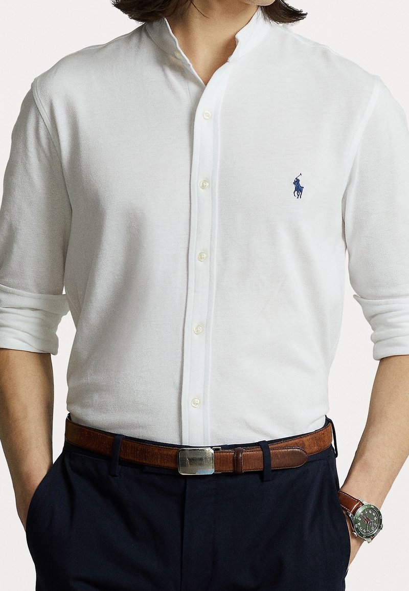 Camisa blanca de botonadura con cuello mao, con un pequeño logotipo azul en el pecho. Combinada con un cinturón de cuero marrón y pantalones azul marino.