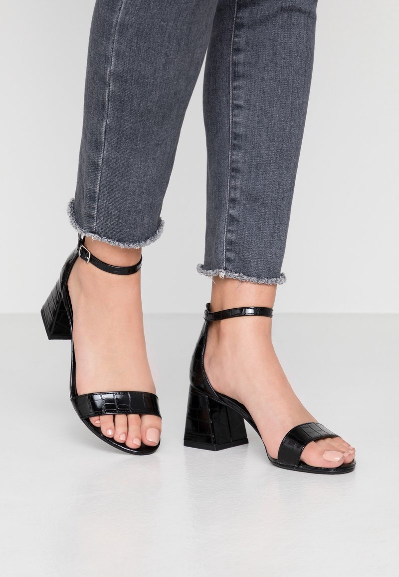 Pieds portant des sandales à talon bloc noires à texture crocodile avec des brides à la cheville, associées à un jean gris à ourlet effiloché.