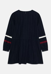 Marineblaues Kleid mit langen Ärmeln und vertikalen Streifen, mit ausgestelltem Rock und rot-weiß gestreiften Akzenten an den Ärmeln. Rückenschlitz mit Knopfverschluss.