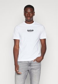 Hvit bomulls t-skjorte med korte ermer, med en svart Calvin Klein-logo i en moderne skrifttype plassert i midten av brystet. Brukt med lysegrå jeans.