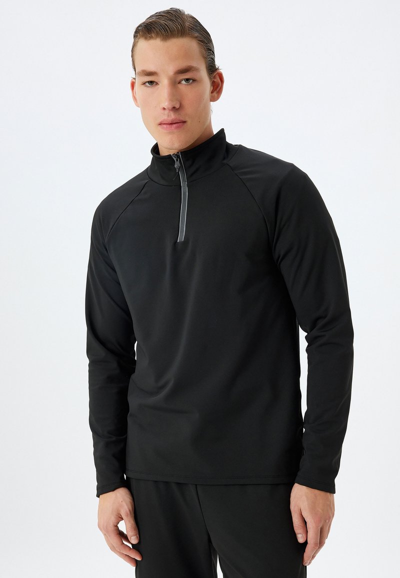 Svart atletisk pullover med krage och halvdragkedja framtill. Tillverkad av ett slätt, stretchigt tyg, har raglanärmar och en smal passform.