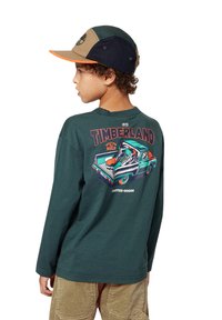 Teal langermet t-skjorte med grafisk trykk av en vintage lastebil og sko, med oransje og hvite detaljer, ribbet halslinning og mykt stoff.
