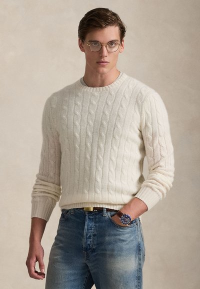 Camisola de malha com padrão de tranças em creme, combinada com jeans azul claro. Detalhes notáveis incluem óculos redondos e um relógio azul com correia castanha.