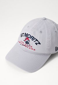 Gorra de béisbol gris con texto bordado "ST. MORITZ" y un gráfico de esquiador. El diseño incluye acentos en rojo y azul marino con un visera curvada.