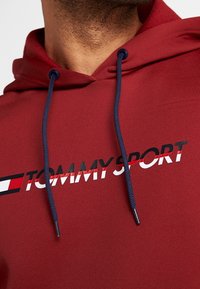 Czerwona bluza z kapturem z granatowymi sznurkami, z logo "TOMMY SPORT" w czerni i bieli oraz czerwoną linią poniżej. Miękka tekstura.