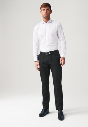 Muž stojaci oblečený v bielej košeli, čiernych nohaviciach s opaskom a čiernych spoločenských topánkach na jednoduchom svetlom pozadí.