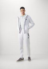 Felpa bianca con zip e accenti neri, polo azzurra chiara, pantaloni della tuta bianchi e sneakers grigie. Presenta loghi sulla felpa e sui pantaloni.