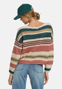 Maglione a righe in tonalità di verde, ruggine, crema e beige. Vestibilità ampia con scollo largo e polsini a costine. Abbinato a jeans blu.